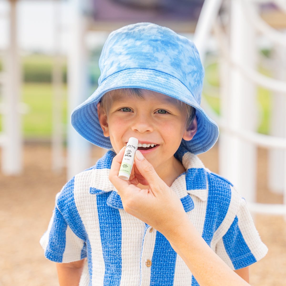 Child wearing a blue hat and striped shirt outdoors, with a mama's hand applying spf lip balm.